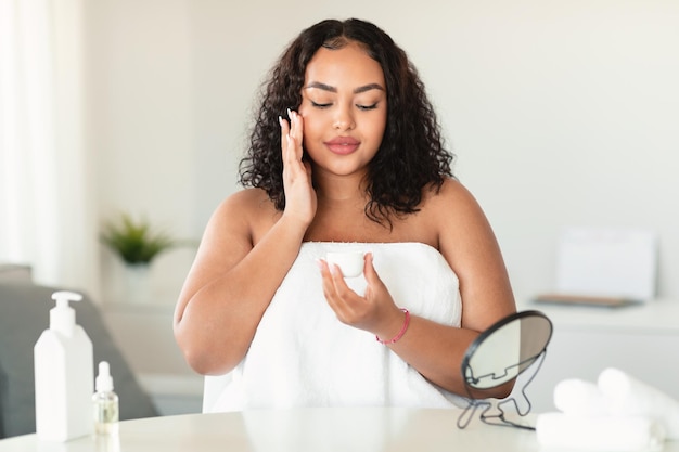 Concepto de cuidado de la piel facial Joven mujer negra con sobrepeso aplicando crema hidratante sosteniendo un tarro de crema sentado en el dormitorio