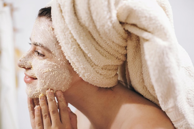 Foto concepto de cuidado de la piel joven mujer feliz en toalla haciendo masaje facial con exfoliante facial orgánico de cerca en un elegante baño chica aplicando crema exfoliante y limpiando la piel