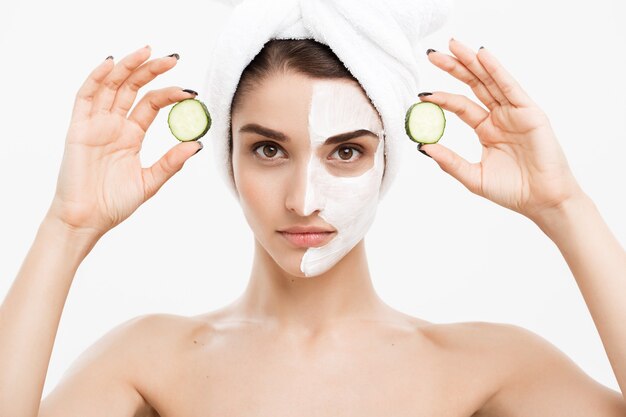 Concepto de cuidado de la piel de la juventud de la belleza - retrato Hermosa mujer caucásica aplicar crema y sosteniendo pepino fresco delante de su cara. Aislado sobre la pared blanca.