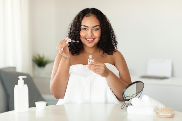 Concepto de cuidado de la piel Mujer negra de talla grande que aplica suero facial con cuentagotas mientras se sienta en casa sonriendo a la cámara