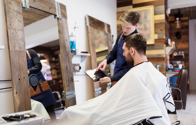 Concepto de cuidado, tecnología y personas: peluquero o barbero mostrando una tableta al hombre en la barbería