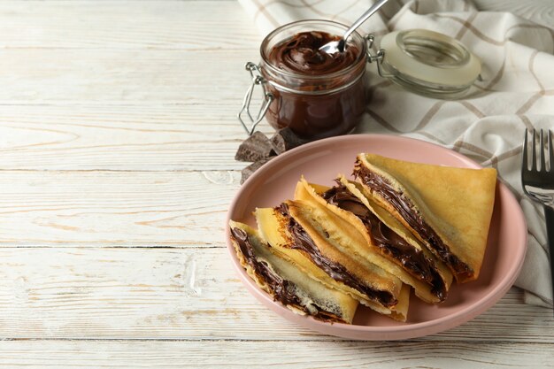 Concepto de delicioso desayuno con crepes con pasta de chocolate sobre fondo blanco de madera