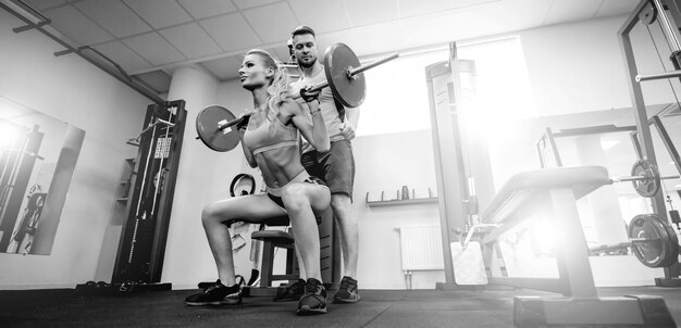 Concepto del deporte, del culturismo, de la forma de vida y de la gente - hombre joven y mujer con la barra que flexiona los músculos y que hace que la prensa del hombro se ponga en cuclillas en gimnasio. posición de rodillas apoyada, trotar en cuclillas.