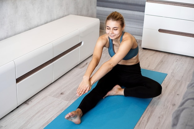 Concepto de deporte, entrenamiento y estilo de vida - mujer estirando la pierna en la estera de yoga azul en casa en la sala de estar