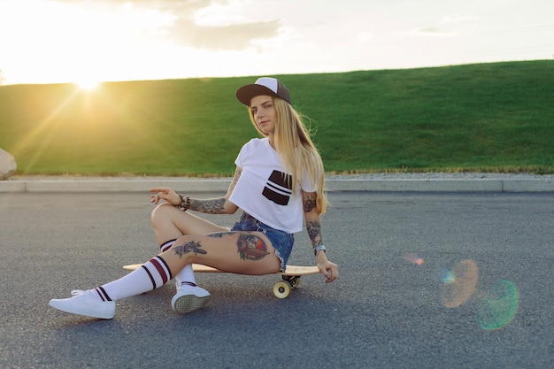 Concepto de deporte, estilo de vida, extremo y personas: tatuajes de hermosas chicas montando longboard en la carretera de la ciudad en un clima soleado. Retrato chica hipster sonriendo con un longboard al atardecer.