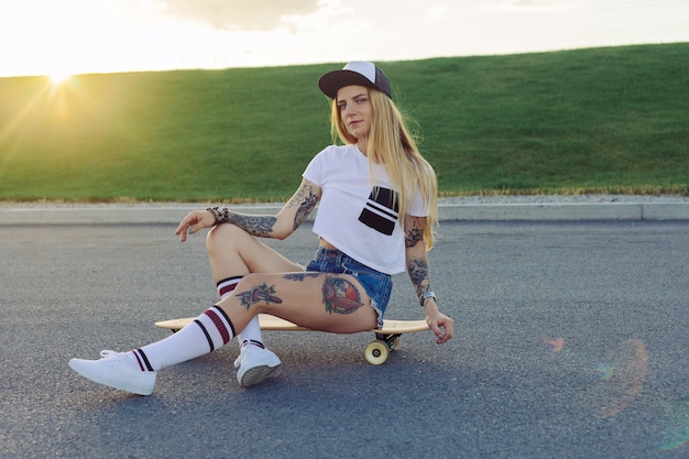 Concepto de deporte, estilo de vida, extremo y personas: tatuajes de hermosas chicas montando longboard en la carretera de la ciudad en un clima soleado. Retrato chica hipster sonriendo con un longboard al atardecer.