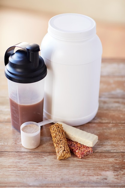 concepto de deporte, fitness, dieta y comida: cierre de frasco, botella de batido de proteínas y barras de muesli en una mesa de madera