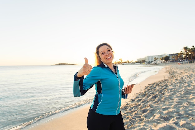 Concepto de deporte, fitness, estilo de vida saludable y funcionamiento - Mujer deportiva motivada haciendo pulgares arriba gesto de éxito después de hacer ejercicio al aire libre en la playa.