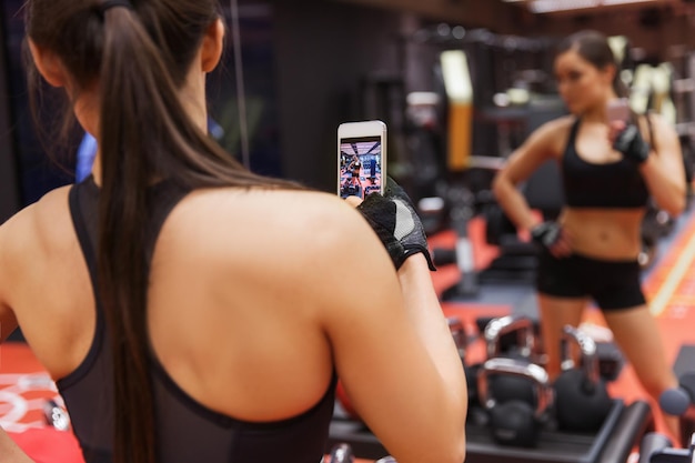 Concepto de deporte, fitness, estilo de vida, tecnología y personas - mujer joven con smartphone tomando selfie espejo en el gimnasio