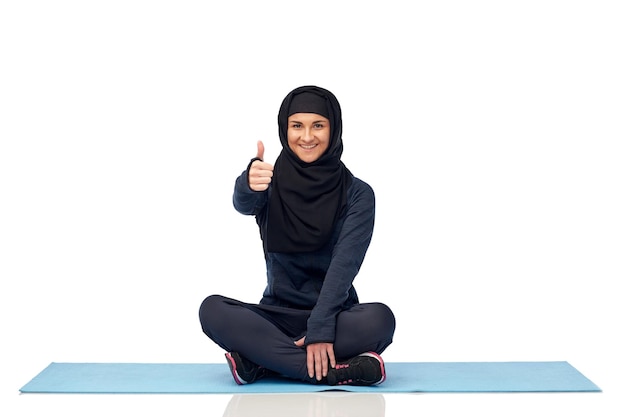 concepto de deporte, fitness y gente - feliz mujer musulmana sonriente en hijab sentada en la alfombra y mostrando los pulgares hacia arriba