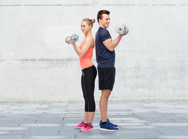 Foto concepto de deporte, fitness y personas: feliz hombre y mujer deportivo con pesas flexionando los músculos sobre fondo de pared de hormigón