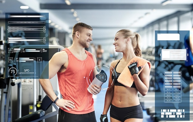 concepto de deporte, fitness y personas - hombre y mujer sonrientes con botella de batido de proteínas y toalla hablando en el gimnasio sobre gráficos virtuales