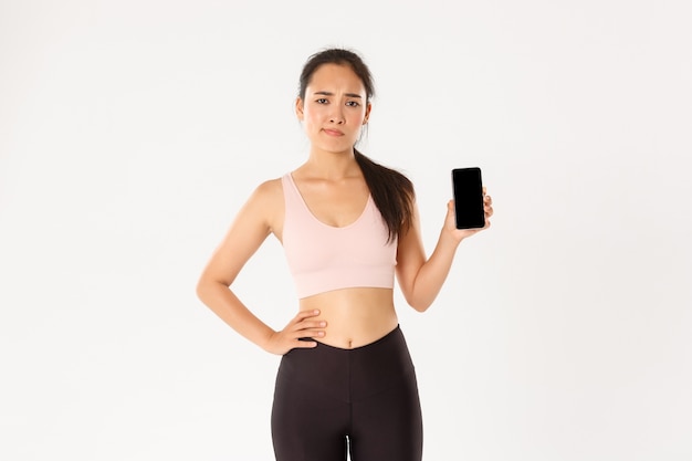 Concepto de deporte, tecnología y estilo de vida activo. Atleta asiática insatisfecha y escéptica, deportista quejándose de la aplicación de fitness, mostrando la pantalla del teléfono inteligente decepcionada