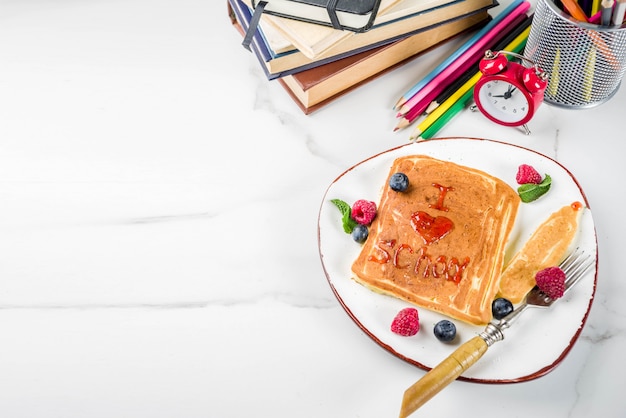 Concepto de desayuno de regreso a la escuela para niños, panqueques con mermelada de frambuesa: me encanta la escuela, en estola de mármol blanco, con libros, reloj despertador, lápices, útiles escolares. Vista superior