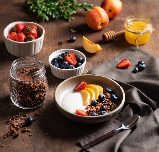 Concepto de un desayuno saludable y nutritivo Delicioso yogur natural con granola casera, melocotón, fresa, miel y arándanos en un bol sobre un fondo de madera oscura Comida dietética