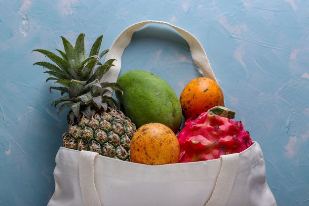 Concepto de desperdicio cero, bolsa textil de compras con frutas tropicales frescas: mango, piña, dragón y maracuyá sobre fondo azul claro, orientación horizontal