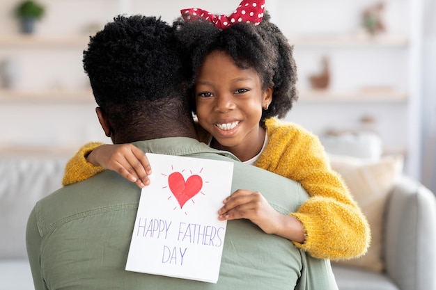 Concepto del día del padre hija negra alegre abrazando a papá y sosteniendo la tarjeta de felicitación