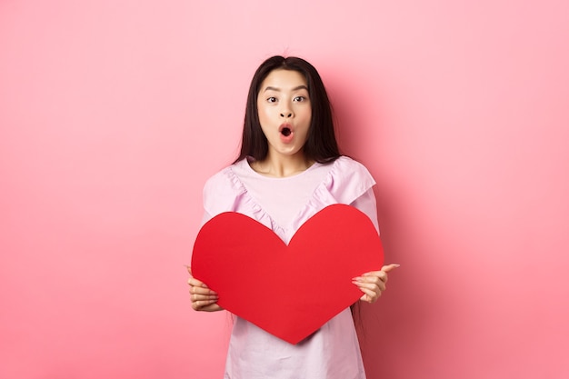 Concepto de día de San Valentín. Chica adolescente asiática sorprendida dice wow, mostrando una gran tarjeta de corazón rojo, recibe un regalo de un admirador o amante secreto, de pie con un vestido romántico sobre fondo rosa.