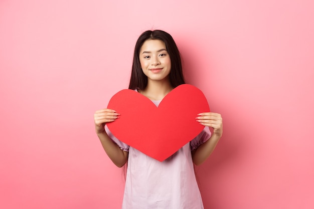 Concepto de día de San Valentín. Linda chica adolescente asiática mostrando una gran tarjeta de corazón rojo, enamorarse, ir a una cita romántica en vestido, sonriendo tiernamente a la cámara, fondo rosa.