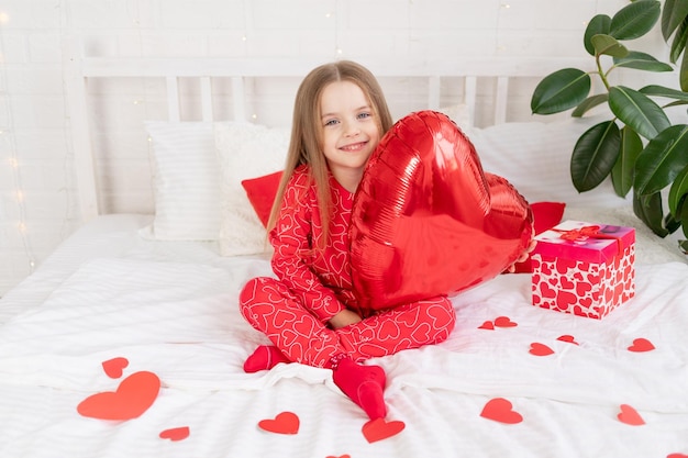 El concepto del día de San Valentín una niña dulce en una cama blanca en casa en pijama rojo entre corazones sosteniendo un gran corazón de globo y sonriendo con felicidad felicitando por las vacaciones