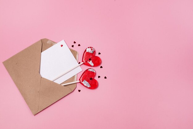 Concepto de día de San Valentín simulado Dos confeti rojo en forma de corazón de piruleta roja y sobre postal artesanal sobre un fondo rosa Espacio de copia mínimo