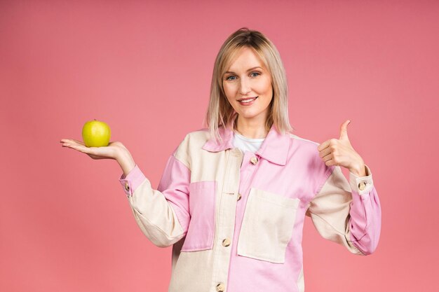 Concepto de dieta de alimentación saludable. Retrato de una joven rubia alegre sosteniendo y comiendo manzana verde aislada de fondo rosa.