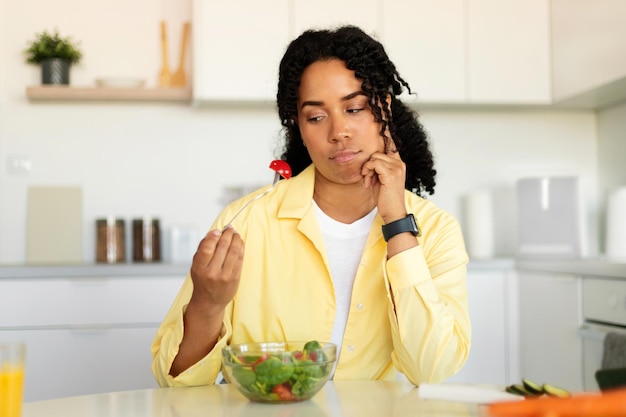 Concepto de dieta Mujer afroamericana infeliz sosteniendo un tenedor con tomate comiendo ensalada de verduras en el interior de la cocina