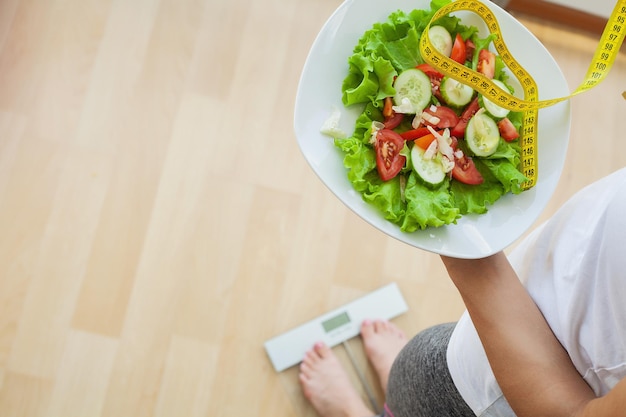 Concepto de dieta, la mujer mide el peso en balanzas electrónicas y ensalada de dieta con cinta métrica amarilla.