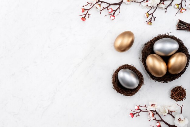 Concepto de diseño de huevos de Pascua dorados y plateados en el nido con flor de ciruelo blanco sobre fondo de mesa de mármol blanco brillante.