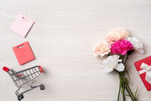 Concepto de diseño de saludo del día de la madre con flor de clavel, idea de regalo de vacaciones y carrito de compras sobre fondo de madera.