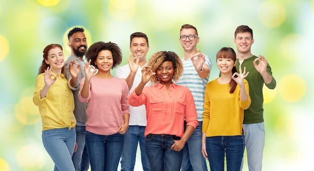 Foto concepto de diversidad, raza, etnia y personas - grupo internacional de hombres y mujeres sonrientes felices que muestran el signo de la mano bien sobre el fondo de las luces de vacaciones verdes