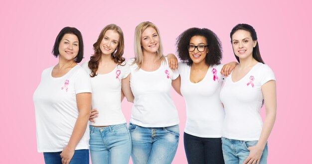 concepto diverso, de salud y de personas - grupo de mujeres felices de diferentes tamaños en camisetas blancas con cinta rosa de concientización sobre el cáncer de mama sobre fondo rosa