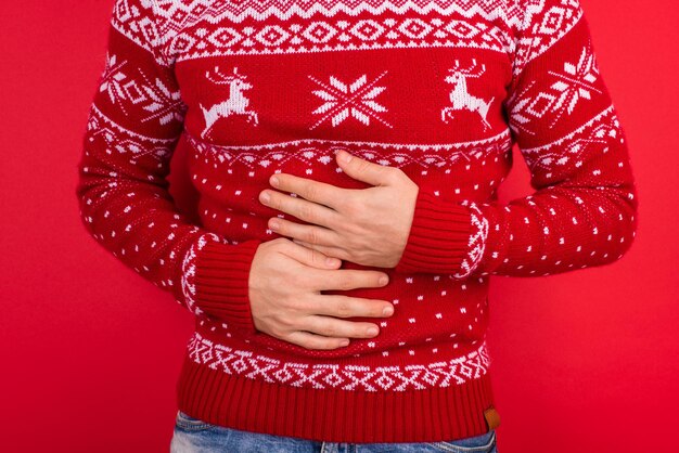 Concepto de dolor de estómago. Cerrar foto de hombres en suéter rojo de invierno con ciervos sosteniendo su estómago aislado sobre fondo rojo.