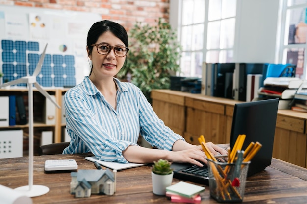 Concepto de ecología y medio ambiente. Mujer de negocios joven que trabaja en una oficina moderna de madera con molino de viento y miniatura 3d en una mesa de madera. arquitecta asiática escribiendo en el diseño de computadoras portátiles.