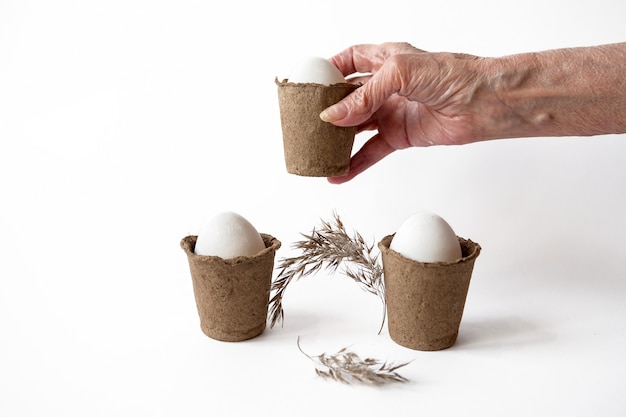 Concepto ecológico con huevos de Pascua blancos en macetas de turba sobre un fondo blanco.