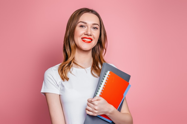 Concepto de educación. Chica estudiante caucásica feliz sonriendo mira cámara y sosteniendo cuadernos, carpetas de libros en manos aisladas sobre fondo rosa studio