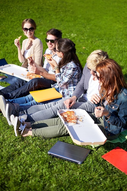 concepto de educación, comida, gente y amistad - grupo de estudiantes adolescentes felices comiendo pizza y sentados en el césped