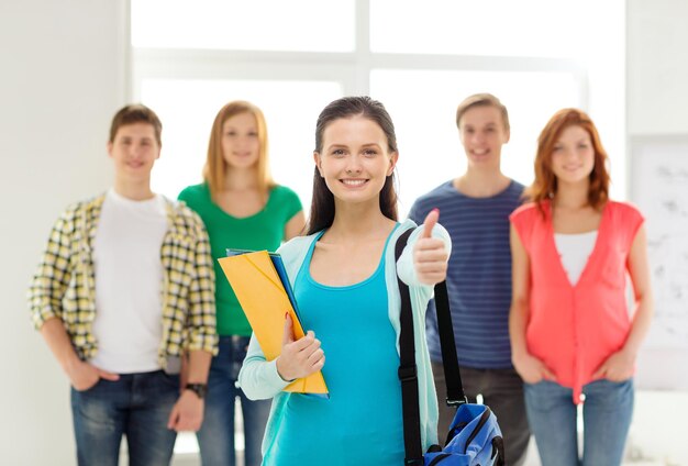 concepto de educación y escuela - grupo de estudiantes sonrientes con una adolescente delante con una bolsa y carpetas mostrando los pulgares hacia arriba