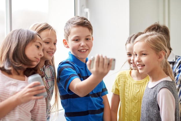 concepto de educación, escuela primaria, bebidas, niños y personas - grupo de escolares con teléfonos inteligentes y latas de refresco tomando selfie en el pasillo