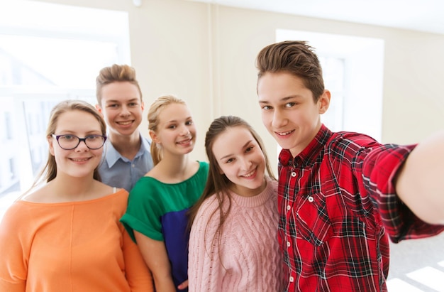 concepto de educación, escuela, tecnología y personas - grupo de estudiantes sonrientes felices tomando fotos con un palo selfie de smartphone en el pasillo