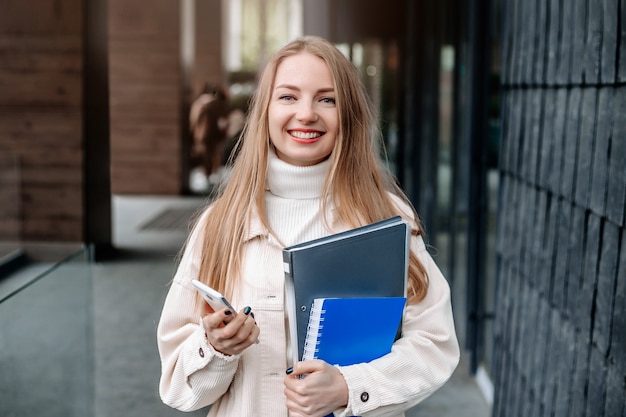 Concepto de educación. Estudiante rubia caucásica sosteniendo un teléfono móvil, carpetas, cuadernos, sonriendo contra el edificio de la Universidad Europea