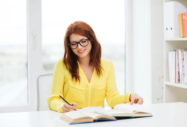 concepto de educación - estudiante sonriente con anteojos leyendo libros y tomando notas en la biblioteca