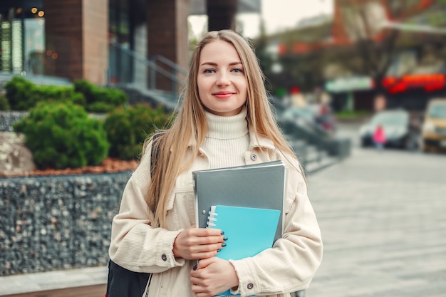 Concepto de educación. Feliz chica estudiante sostiene carpetas cuadernos libros en manos sonríe contra un moderno edificio universitario