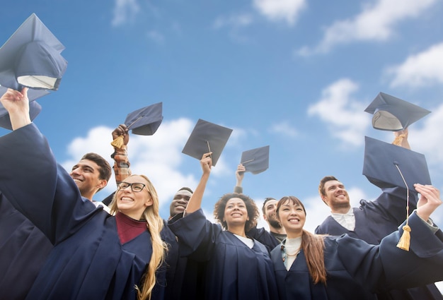 concepto de educación, graduación y personas: grupo de estudiantes internacionales felices con vestidos de soltero agitando tablas de mortero o sombreros sobre el cielo azul y el fondo de las nubes