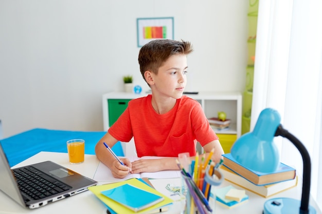concepto de educación, infancia y personas - niño estudiante feliz con computadora portátil escribiendo en un cuaderno en casa