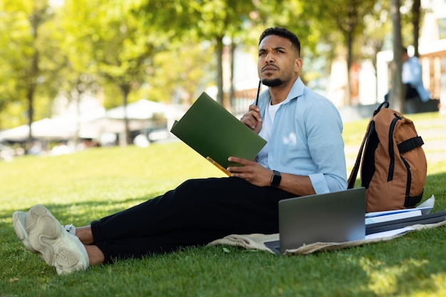 Concepto de educación en línea Estudiante afroamericano pensativo que estudia al aire libre usando una computadora portátil y tomando notas