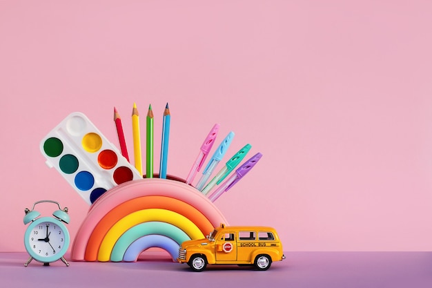 El concepto de educación y regreso a la escuela. Papelería en una caja de lápices, un reloj despertador y un autobús escolar de coche de juguete sobre la mesa sobre un fondo rosa