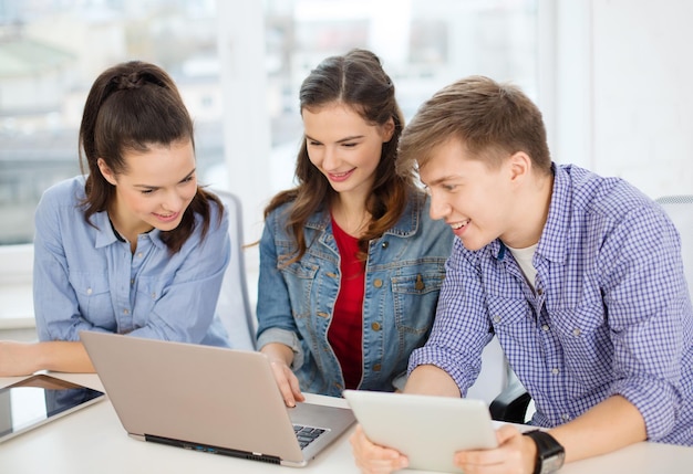 concepto de educación, tecnología, escuela e Internet - tres estudiantes sonrientes con laptop y tablet pc en la escuela