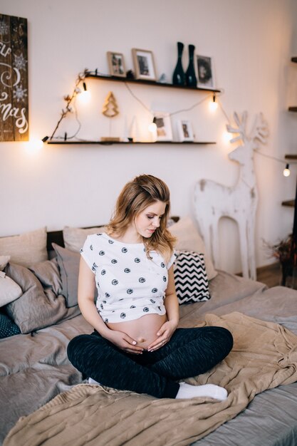 Concepto de embarazo, descanso, personas y expectativa - cerca de la mujer embarazada sonriente feliz sentada en la cama y tocando su vientre en casa