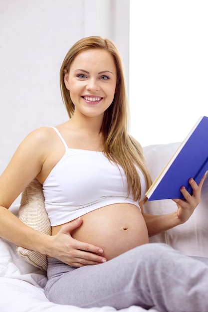 concepto de embarazo y maternidad - mujer embarazada sonriente sentada en un sofá y leyendo un libro en casa